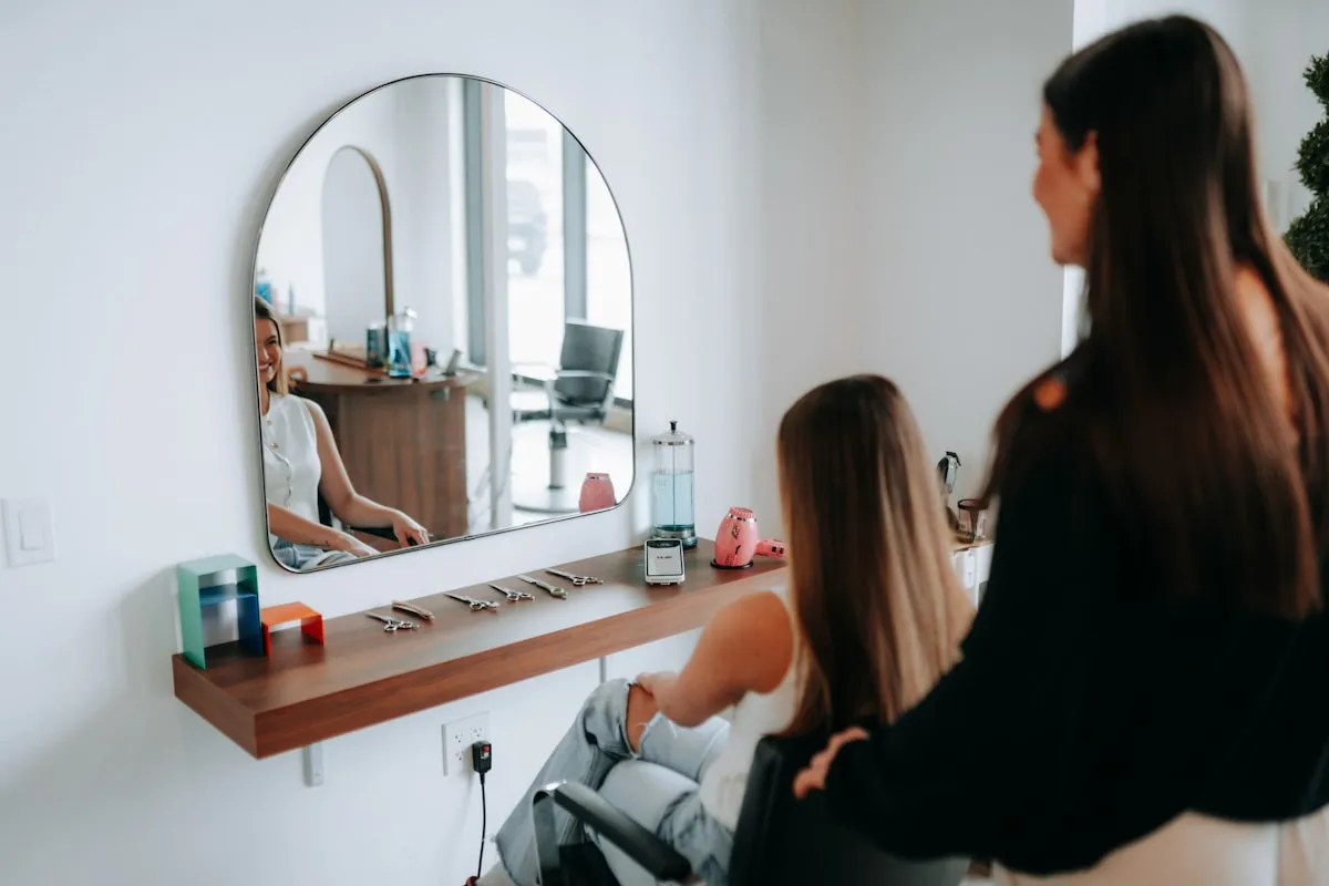Cliente sorrindo para o próprio reflexo no espelho do salão depois do atendimento, com a cabeleireira em pé ao lado
